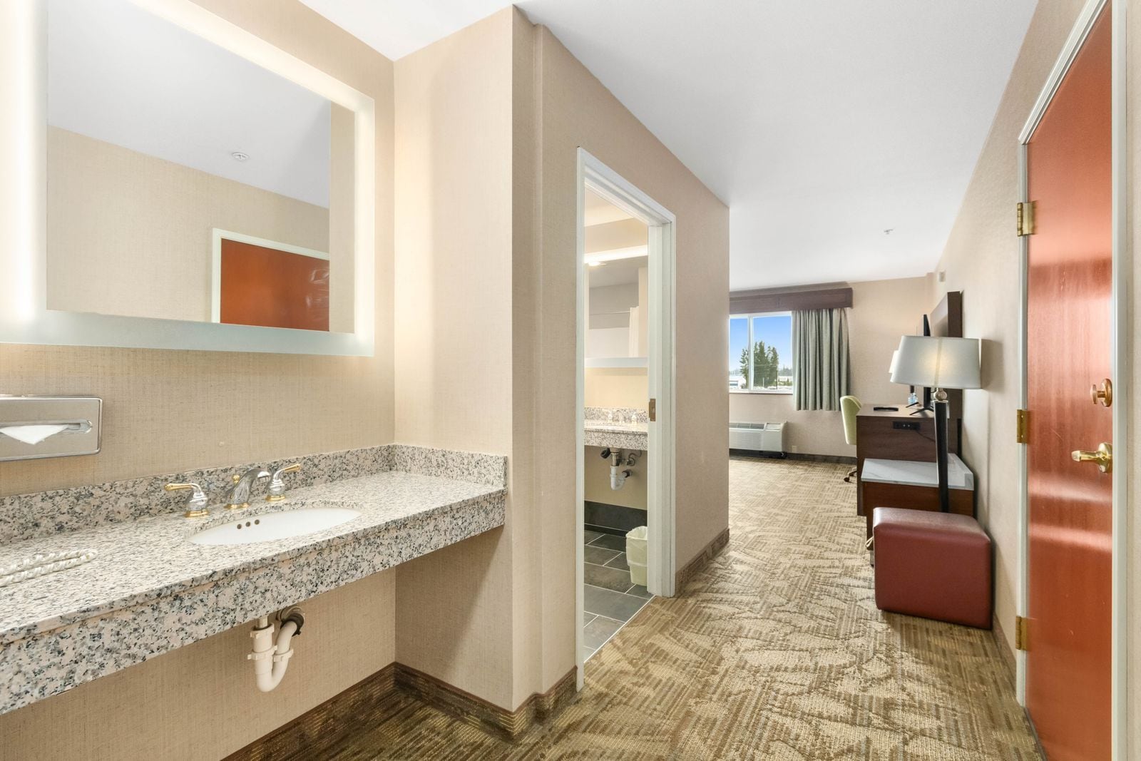 A hotel bathroom with a granite countertop sink, a mirror, and a towel dispenser.