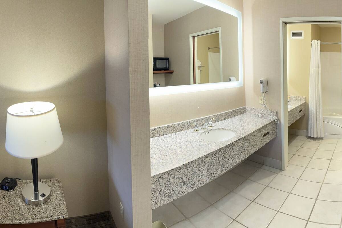 Habitación de hotel con cama, escritorio, silla, televisor, lámpara y baño con ducha y lavamanos.