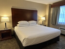 A hotel room with a large bed, two bedside tables with lamps, a headboard, a window with curtains, and an air conditioning unit.