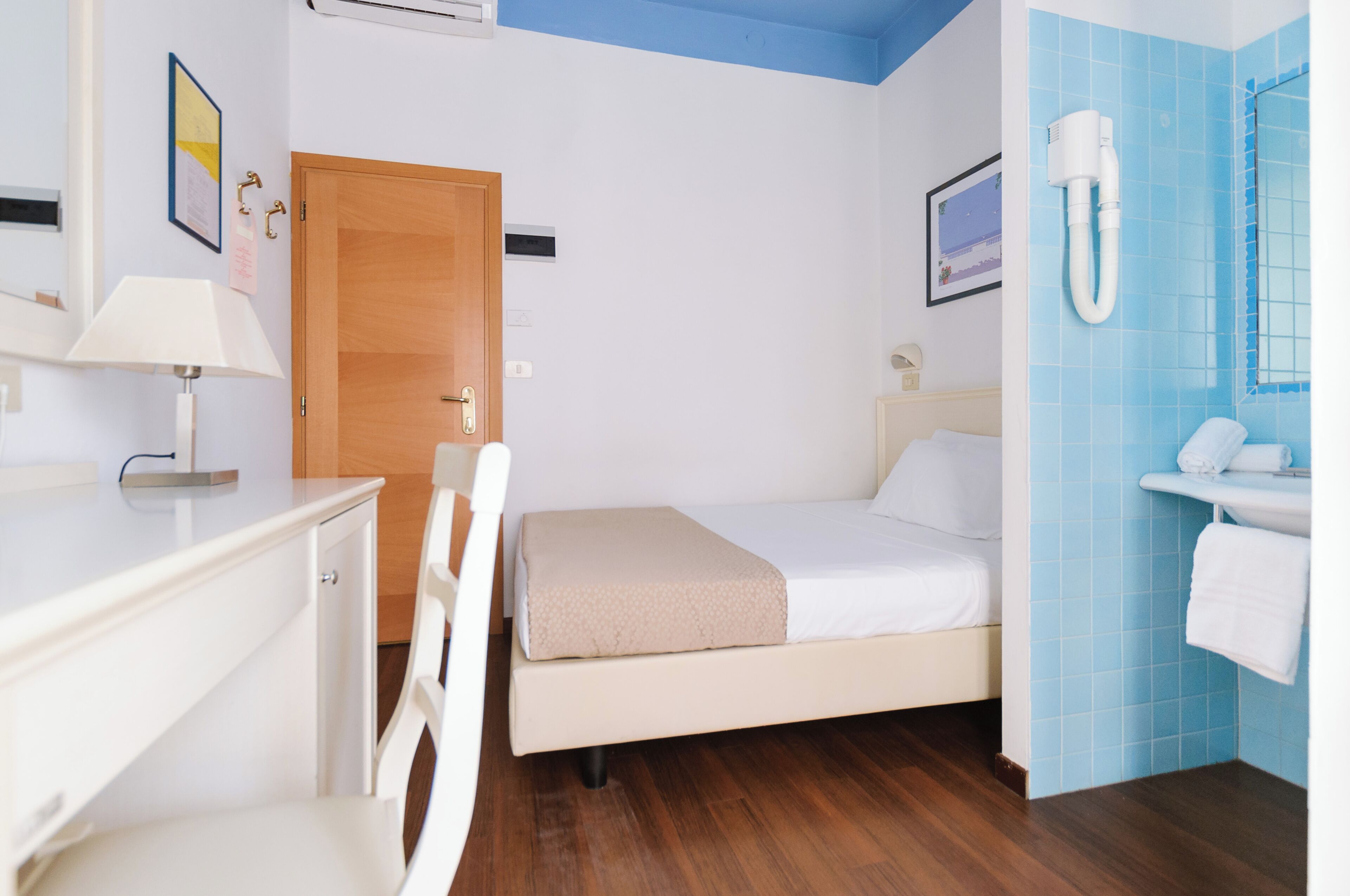 A hotel room with a bed, a desk, a chair, a wooden door, a bathroom with a sink and a mirror, and a wall-mounted air conditioning unit.