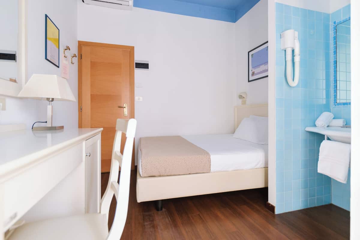 A hotel room with a bed, a desk, a chair, a wooden door, a bathroom with a sink and a mirror, and a wall-mounted air conditioning unit.