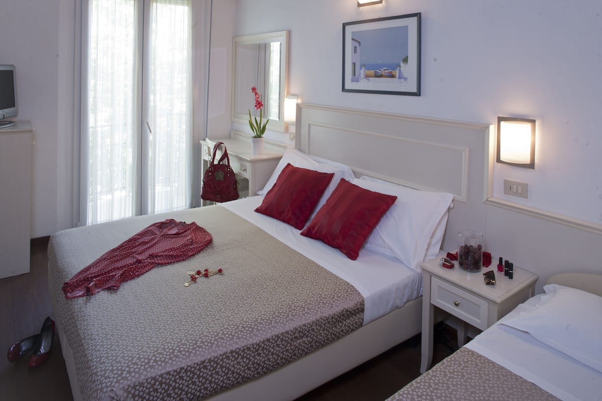 A hotel room with a bed, a bedside table, a nightstand, a lamp, a mirror, and a window with curtains.