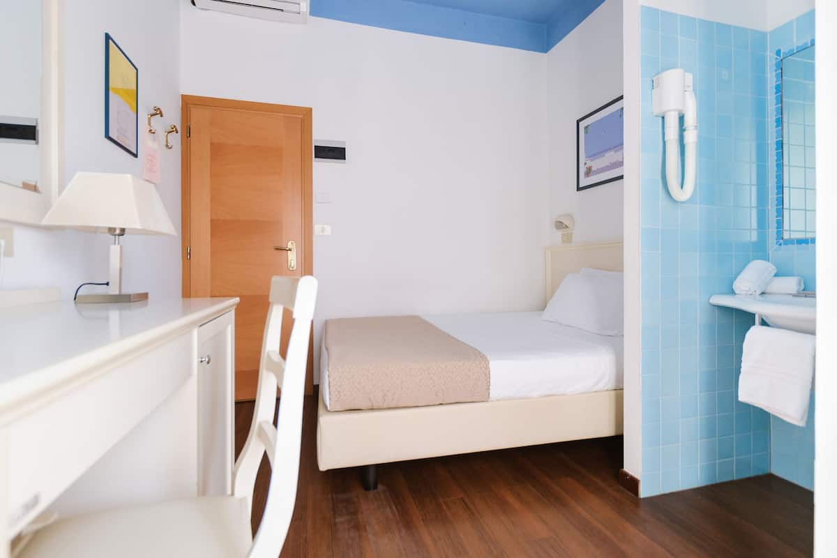 A hotel room with a bed, a desk, a chair, a wooden door, a bathroom with a sink and a mirror, and a wall-mounted air conditioning unit.