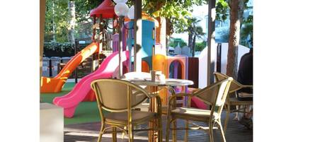 An outdoor seating area with wicker chairs and a table, surrounded by colorful play structures and a slide.