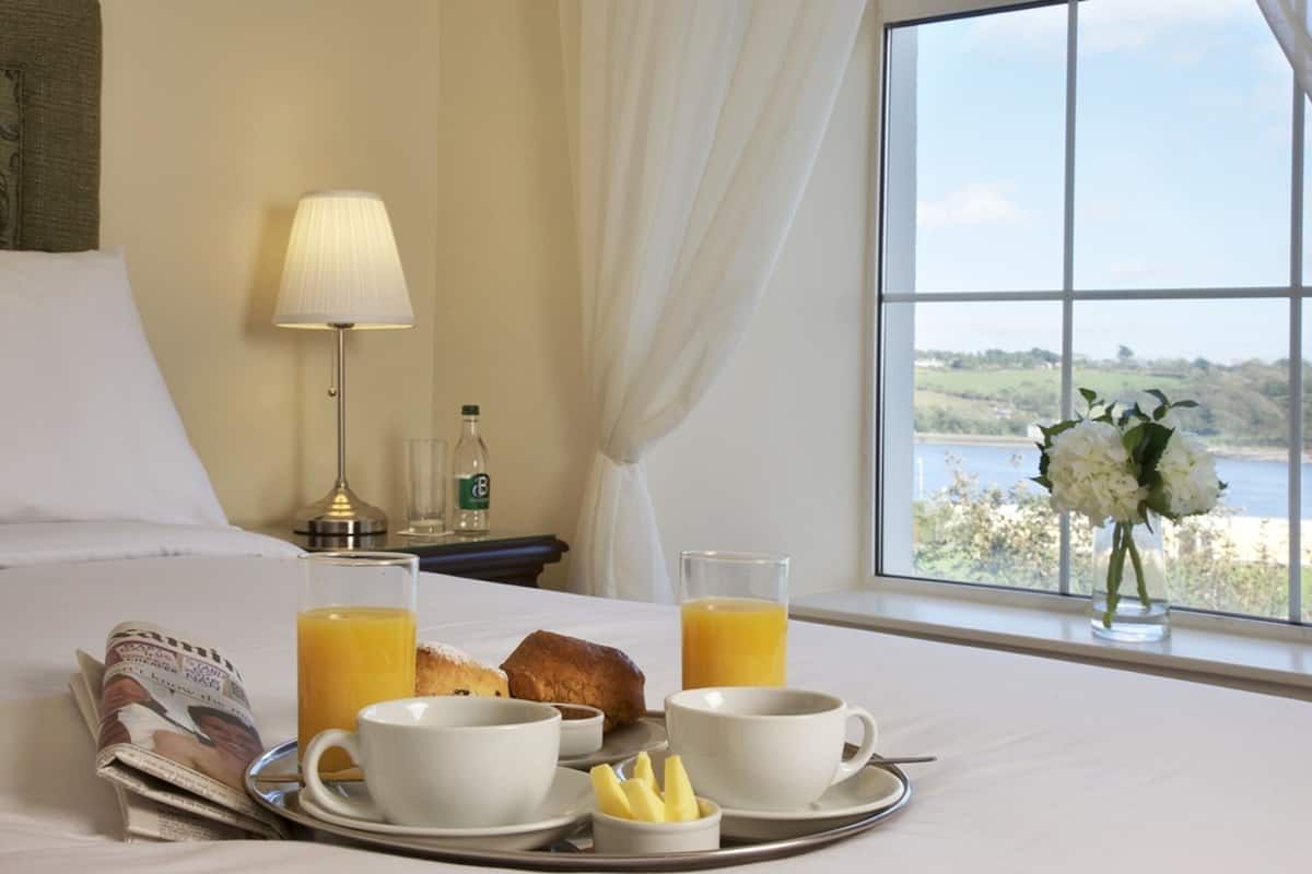 A hotel room with a breakfast tray on the bed, a lamp, a bottle, and a view of the outdoors.