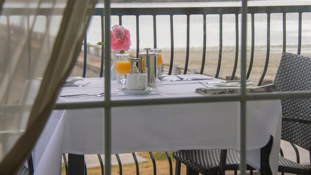 Ein Tisch mit weißer Tischdecke, einer rosa Blume, einem Glas Orangensaft und einer Metallkanne, mit Blick auf einen Strand.