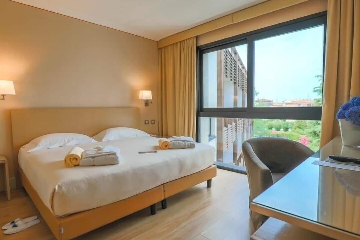 Habitación de hotel con una cama grande, un escritorio y vistas a edificios y zonas verdes.