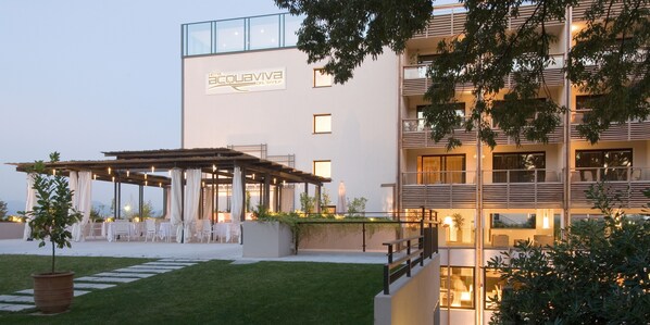 A hotel with a modern design, featuring a covered outdoor seating area with tables and chairs, a well-maintained lawn, and a balcony with potted plants.