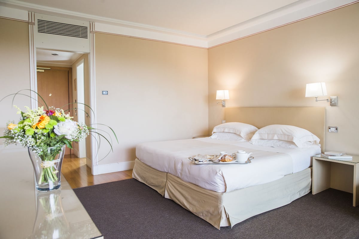 Una habitación de hotel con una cama, una mesita de noche, una lámpara y un florero con flores.