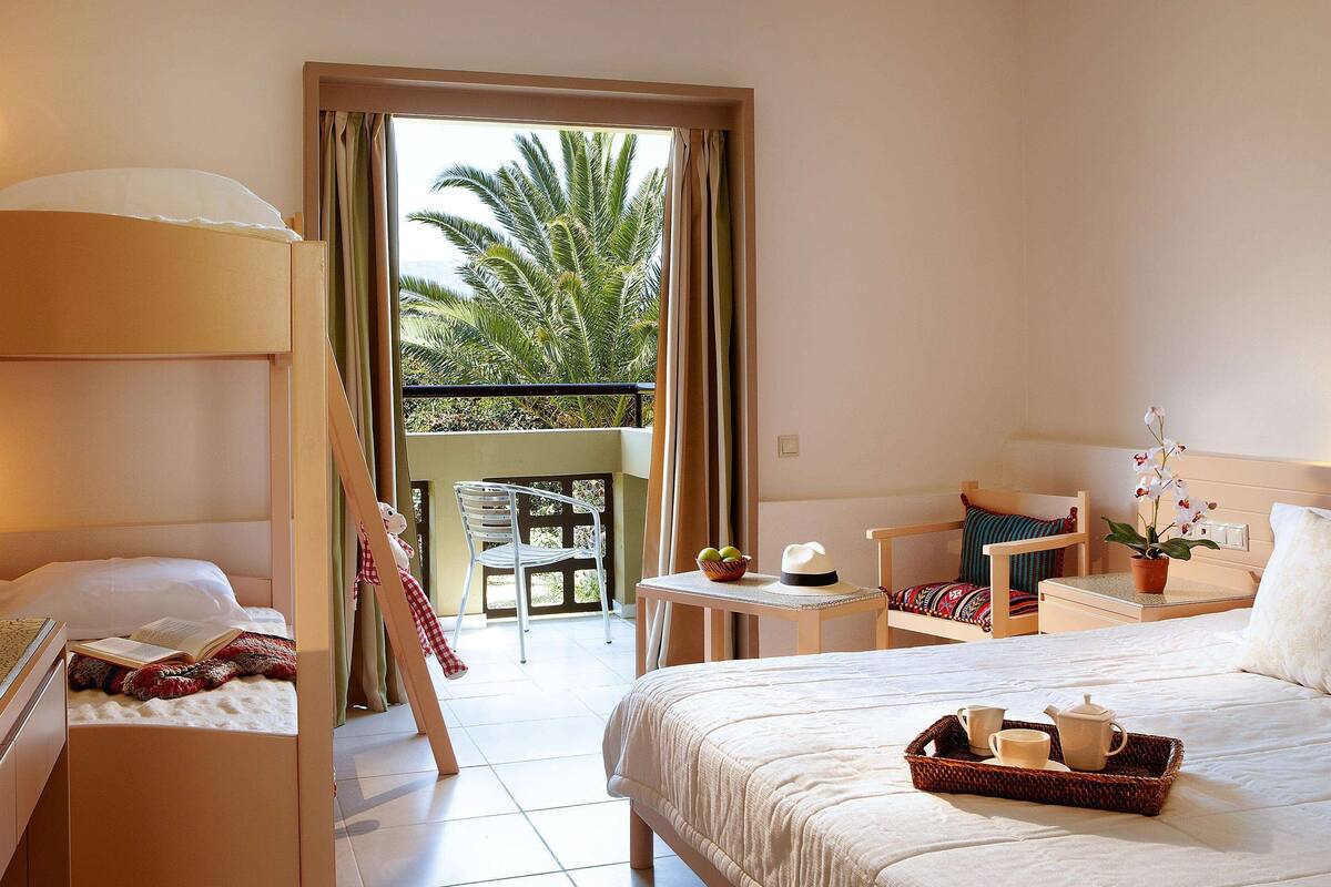 A hotel room with a balcony, a bed with a tray of breakfast items, a chair, a small table with a lamp, and a view of palm trees.