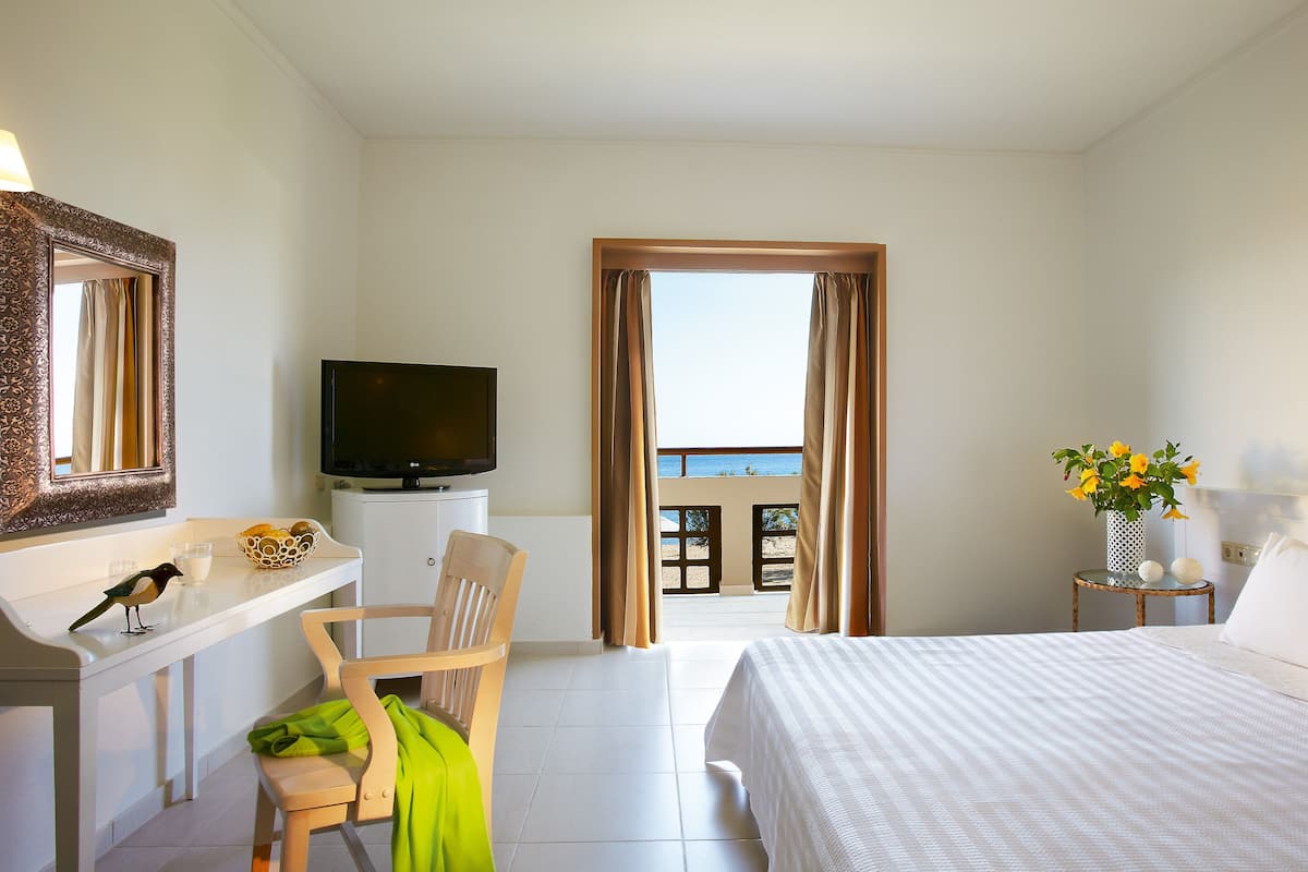 A hotel room with a bed, a desk with a television, a chair, a window with curtains, and a view of the sea.