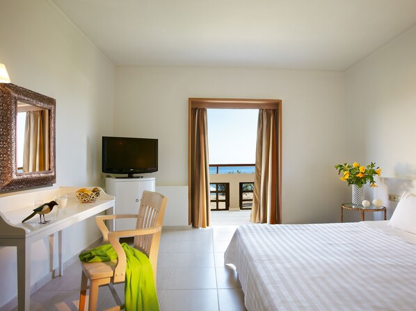 A hotel room with a bed, a desk with a television, a chair, a window with curtains, and a view of the sea. - Grecotel Meli Palace (Agios Nikolaos)