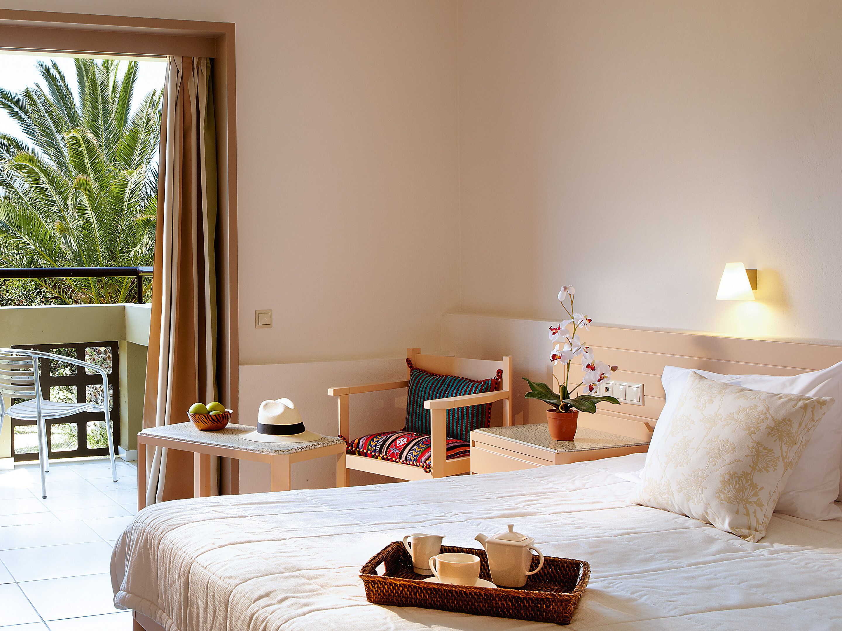 A hotel room with a bed, a small table with a tray of breakfast items, a chair, a potted orchid, and a balcony with a view of palm trees.