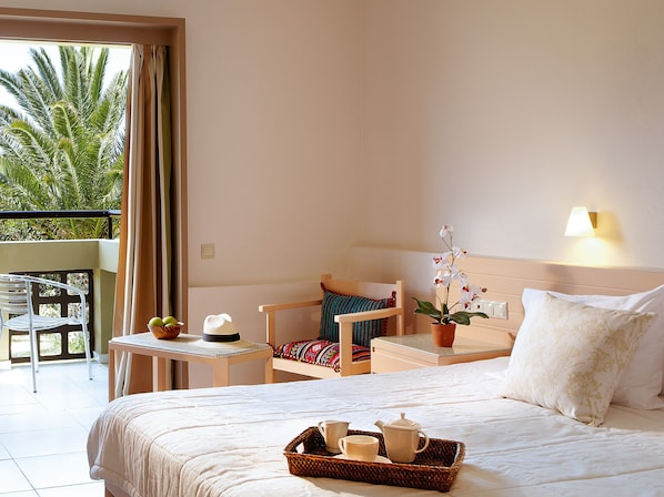 A hotel room with a bed, a small table with a tray of breakfast items, a chair, a potted orchid, and a balcony with a view of palm trees. - Grecotel Meli Palace (Agios Nikolaos)