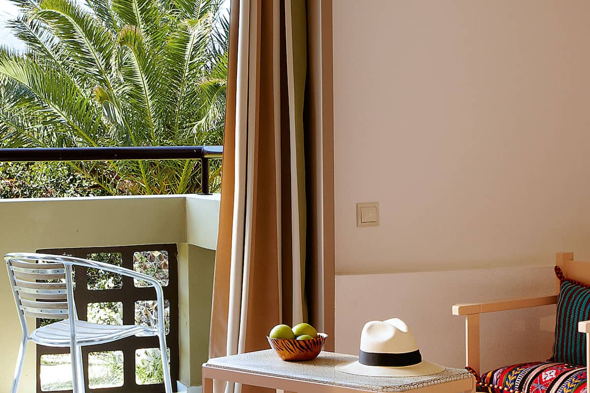 A balcony with a white chair, a small table with a hat and a bowl of fruit, and a view of palm trees.