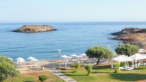 A beach with white umbrellas and lounge chairs, a rocky outcrop in the sea, and a clear sky. - Grecotel Meli Palace (Agios Nikolaos)