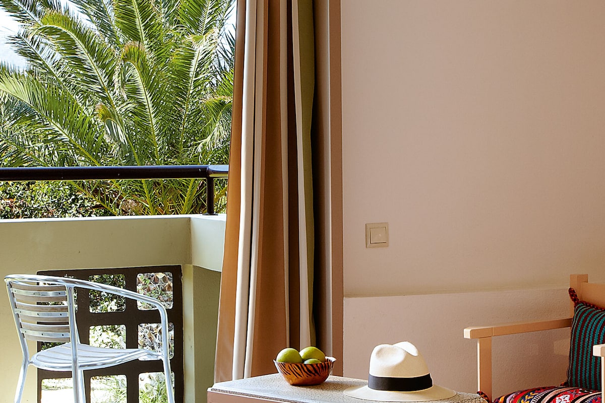 A balcony with a white chair, a small table with a hat and a bowl of fruit, and a view of palm trees.