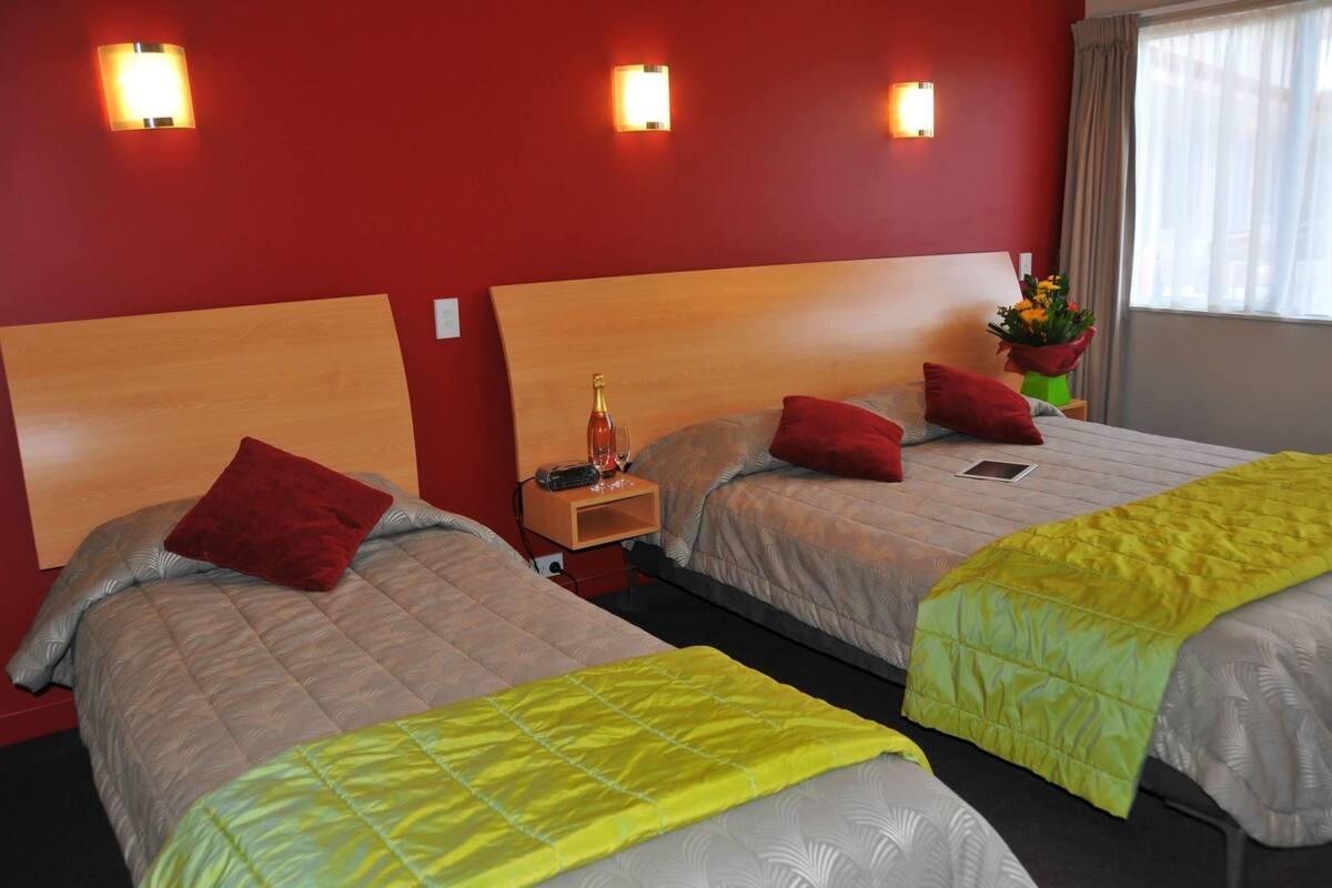 A hotel room with two beds, each with a wooden headboard, red pillows, and a bedside table with a bottle and glasses.