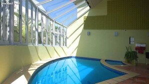 An indoor swimming pool with a skylight, tiled floor, and a view of palm trees outside. - Park View Motor Lodge (Hamilton)