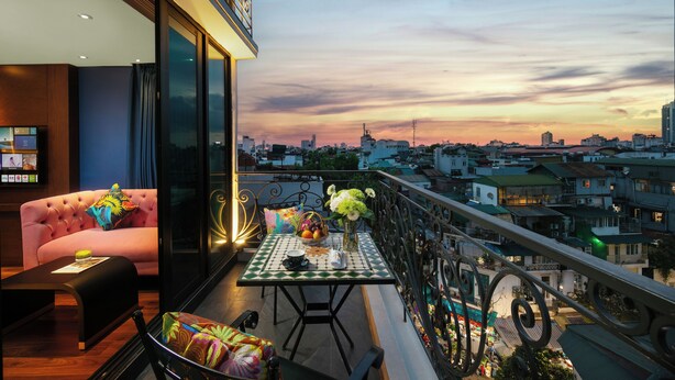 A balcony with a table set for dining, a sofa with colorful cushions, and a cityscape view at sunset. - Aurora Central Hotel  (Hanoi)
