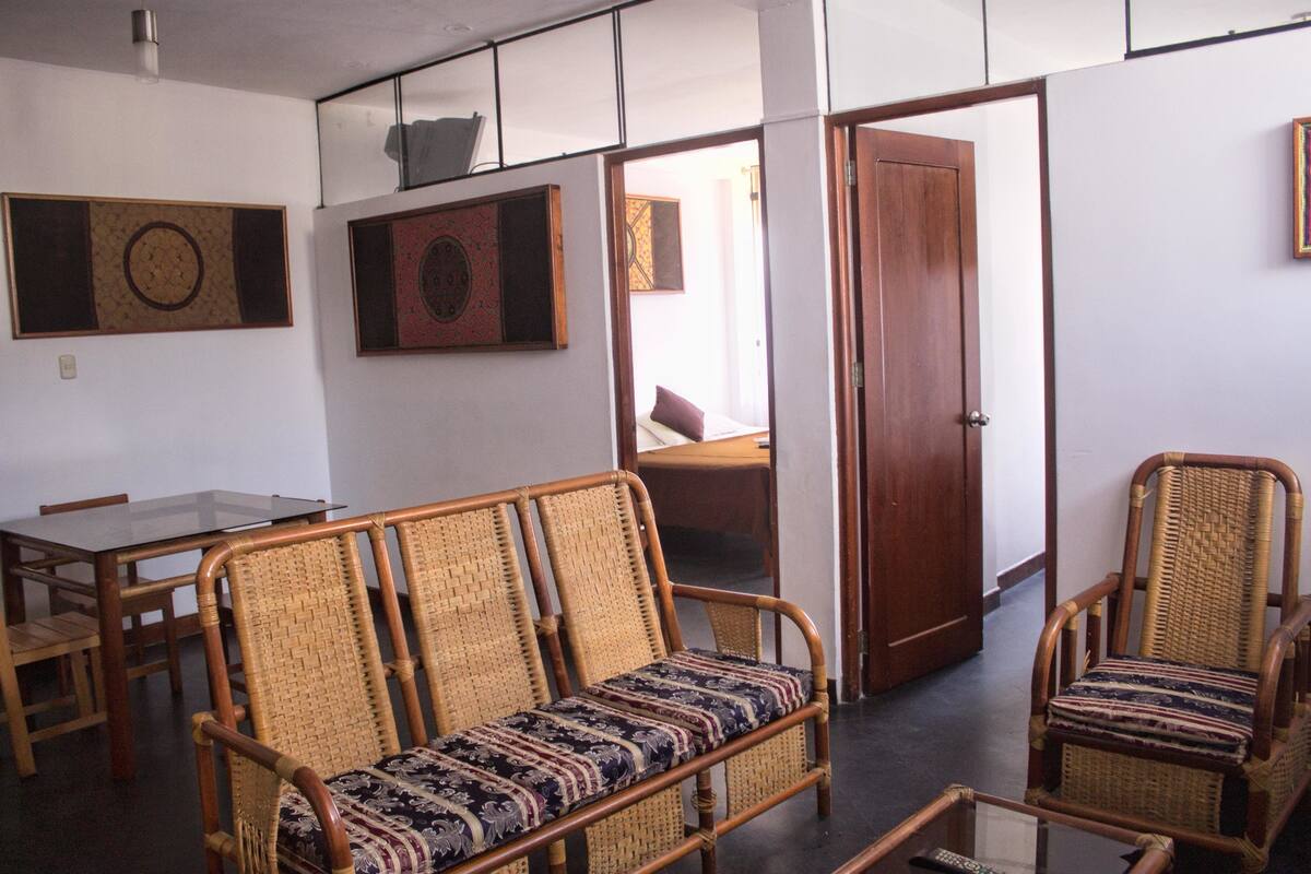 Una habitación con una mesa de comedor, dos sofás de mimbre, una puerta de madera y vista a otra estancia.