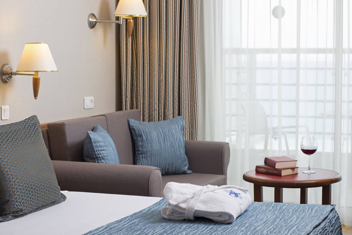 A hotel room with a bed, a sofa, a table with a glass of red wine, and a window with curtains.
