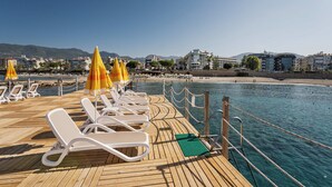 A wooden deck with white sun loungers and yellow umbrellas, overlooking a beach and city buildings. - Grand Kaptan Hotel - Ultra All Inclusive (Alanya)