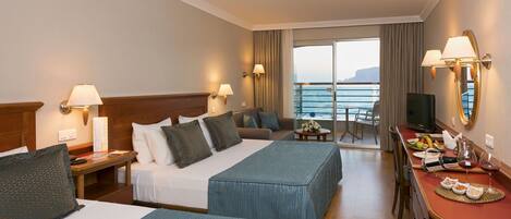 A hotel room with two beds, a desk with a TV, and a balcony with a view of the sea.