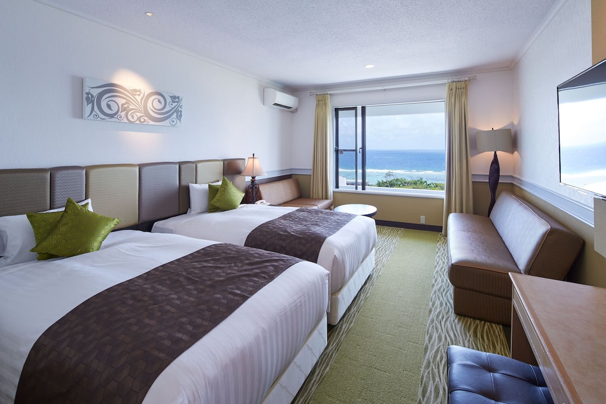 A hotel room with two beds, a seating area, and a large window with a view of the ocean.