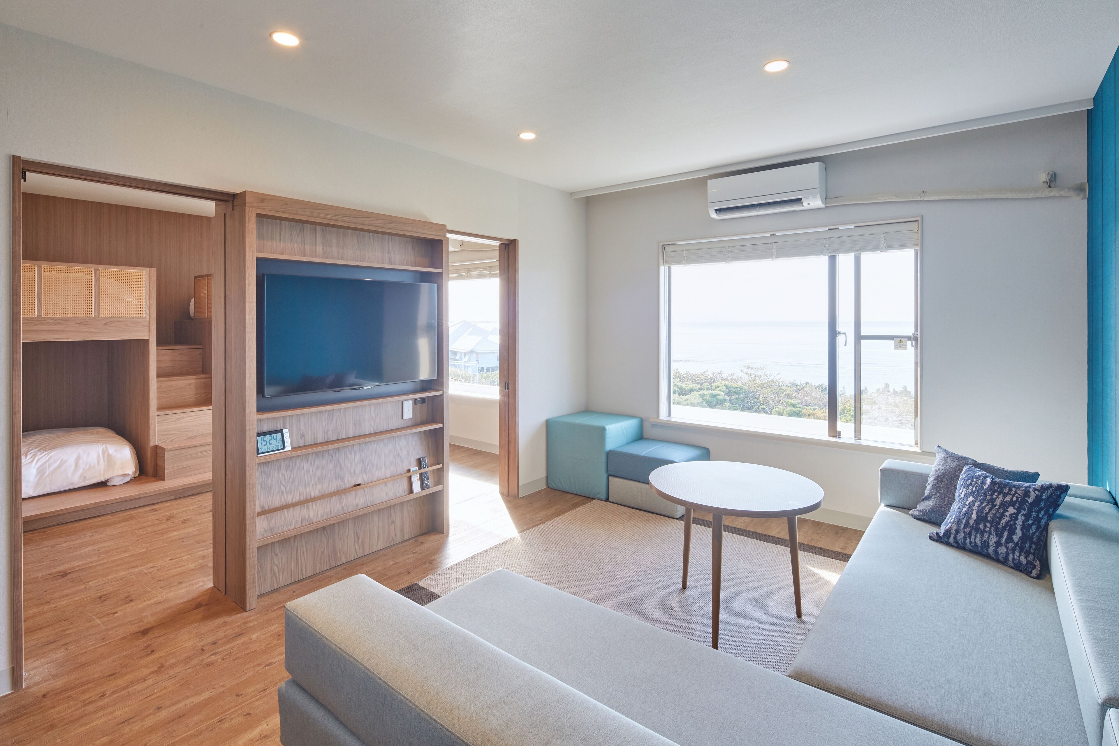 A modern hotel room with a sofa, a small table, a TV, a wardrobe, and a window with a view.