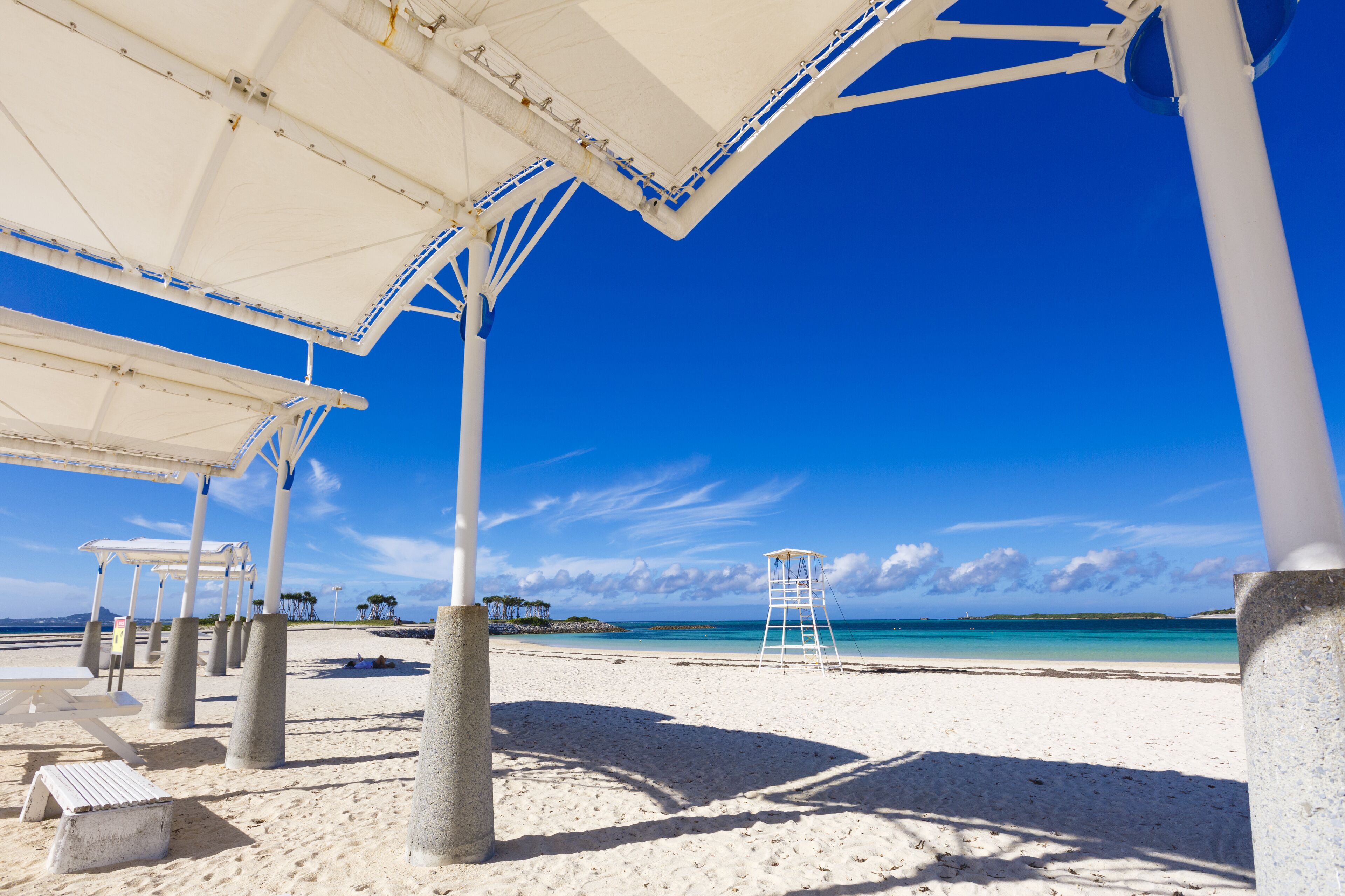 Ein Strand mit einer weißen Sonnenschirmkonstruktion, Betonpfeilern und einem Rettungsschwimmturm.