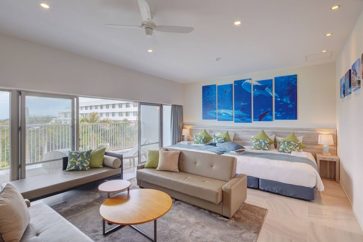 A modern hotel room with a large bed, a sofa, a small table, and a balcony with a view of the city.