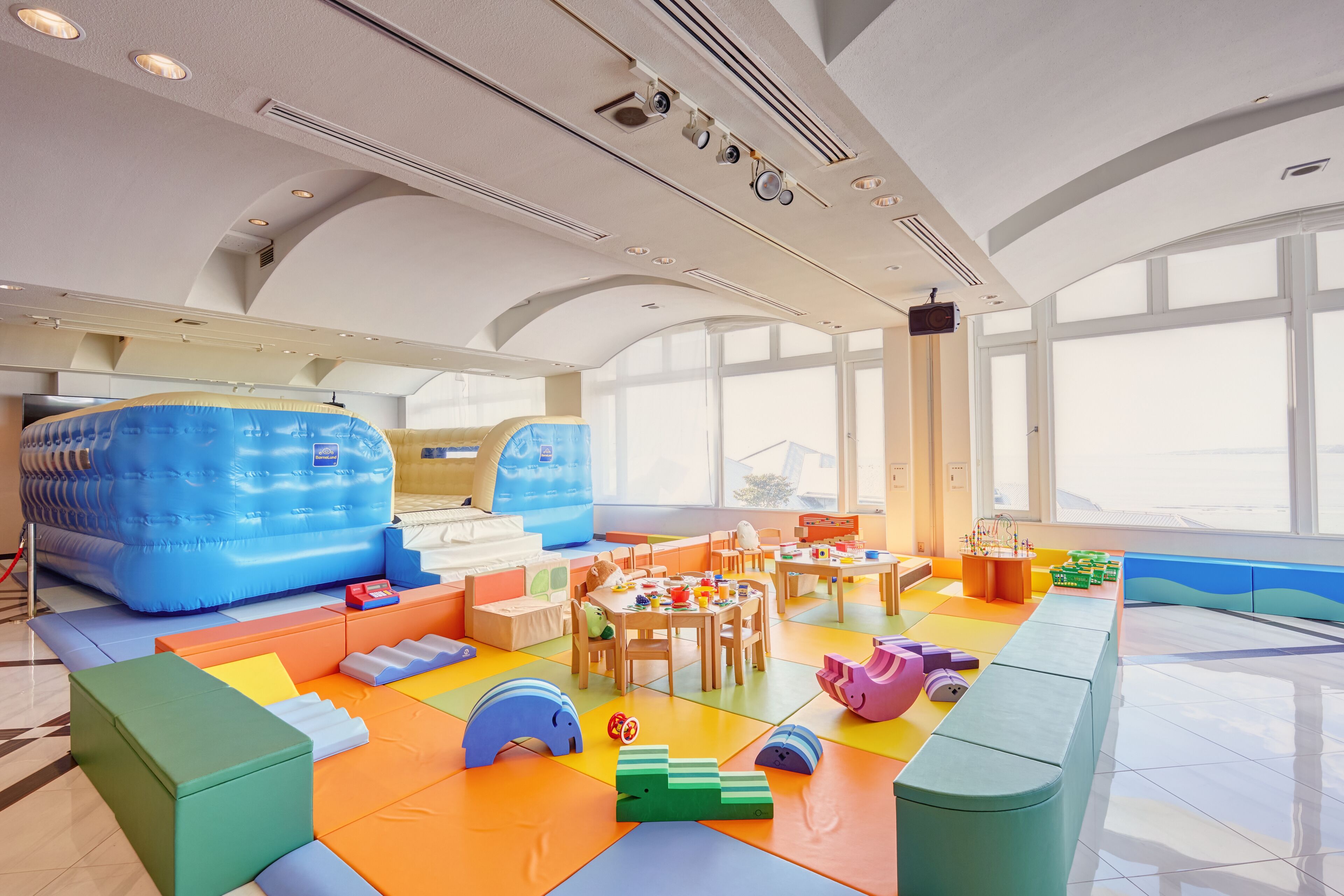 Ein großzügiger Indoor-Spielbereich mit bunten Spielmatten, weichen Spielgeräten und Spieltischen.