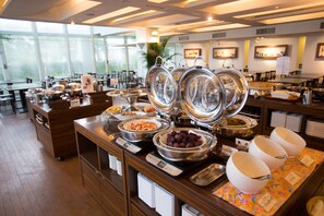 A buffet setup with multiple food stations, including bowls of fruits and dishes, in a well-lit dining area with large windows.