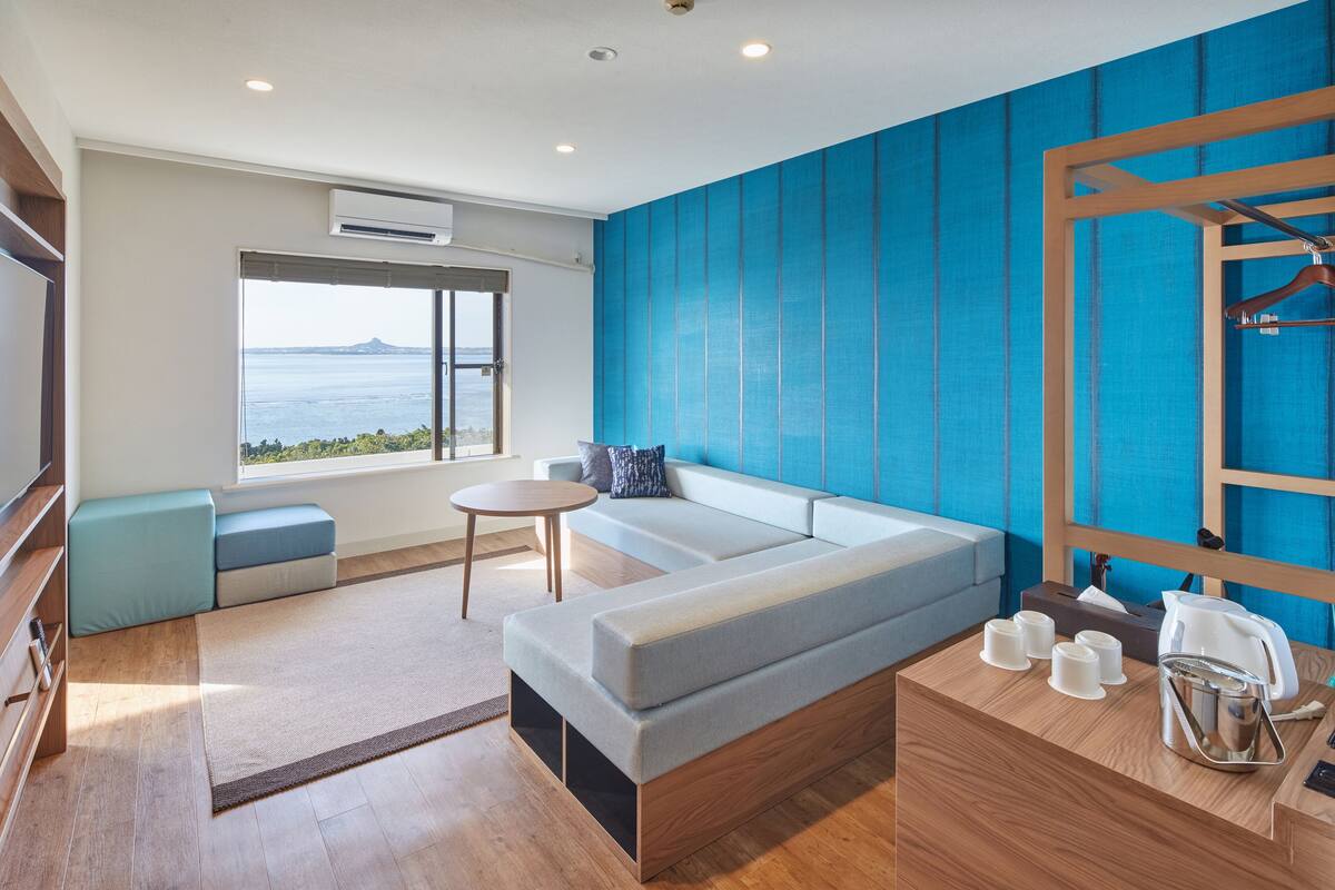 A modern hotel room with a large window, a sofa, a small table, and a wooden desk with a kettle and cups.