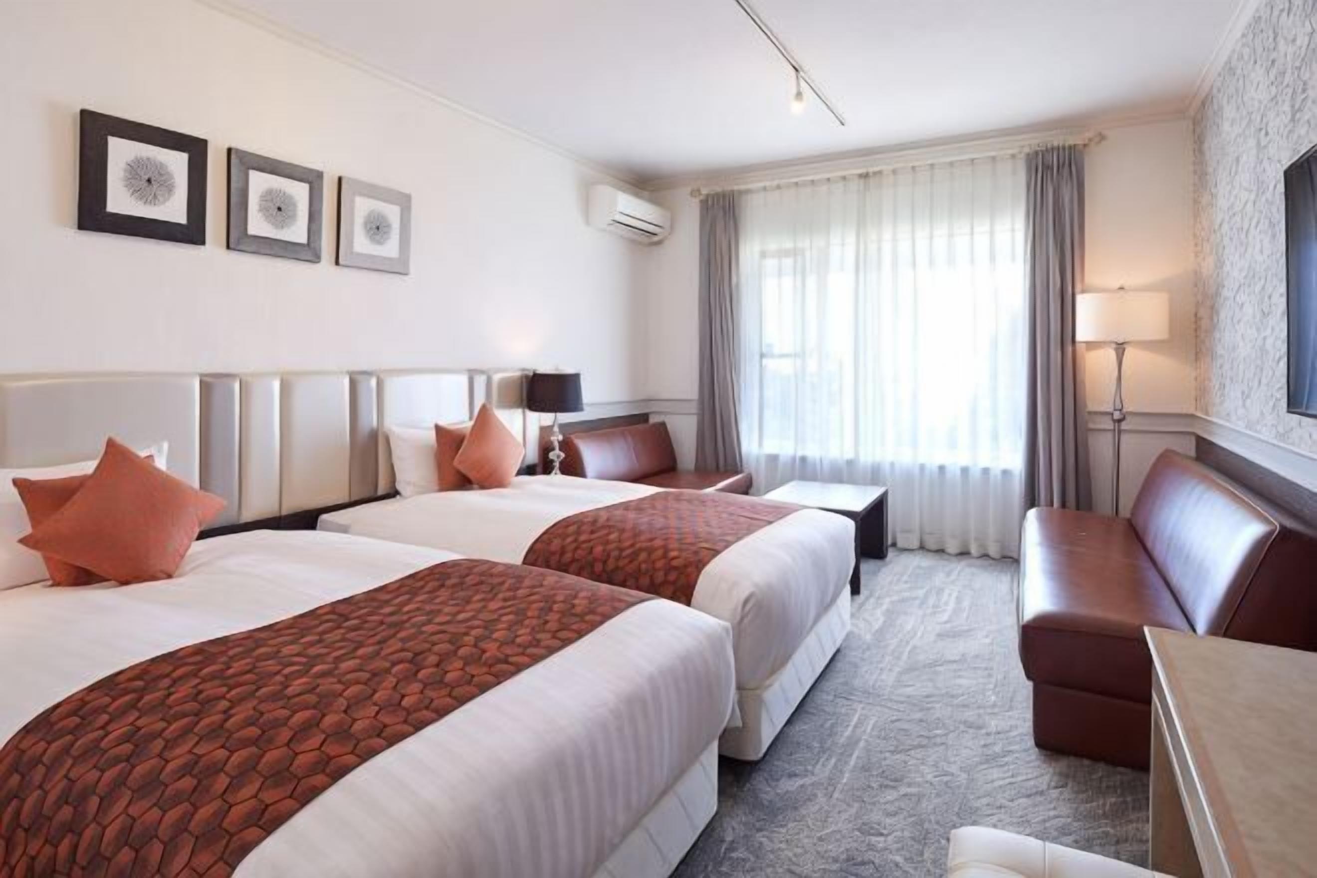 A hotel room with two beds, a sofa, a desk, and a large window with curtains.