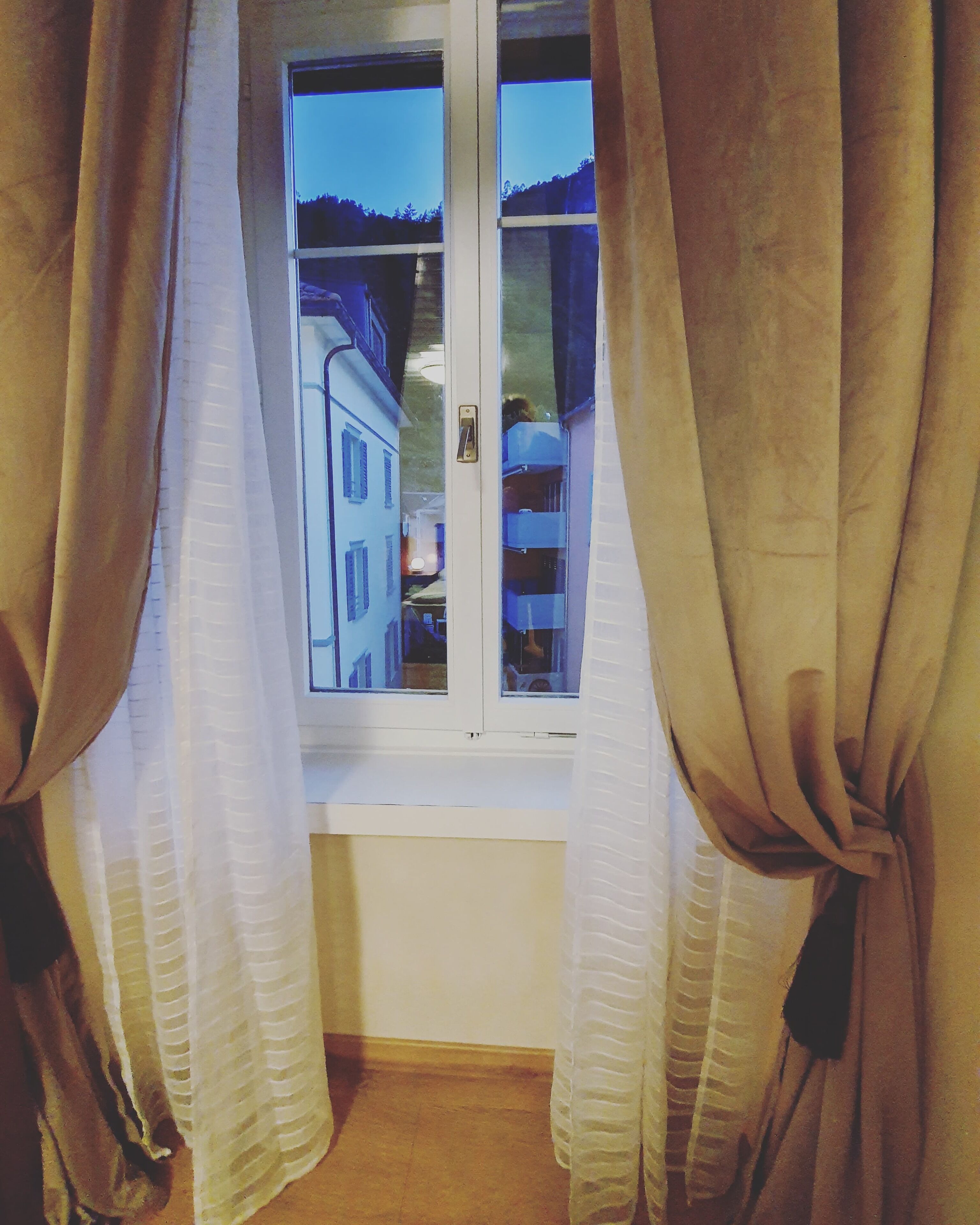 Una ventana con cortinas recogidas, mostrando una vista de un edificio con varias ventanas y una puerta.
