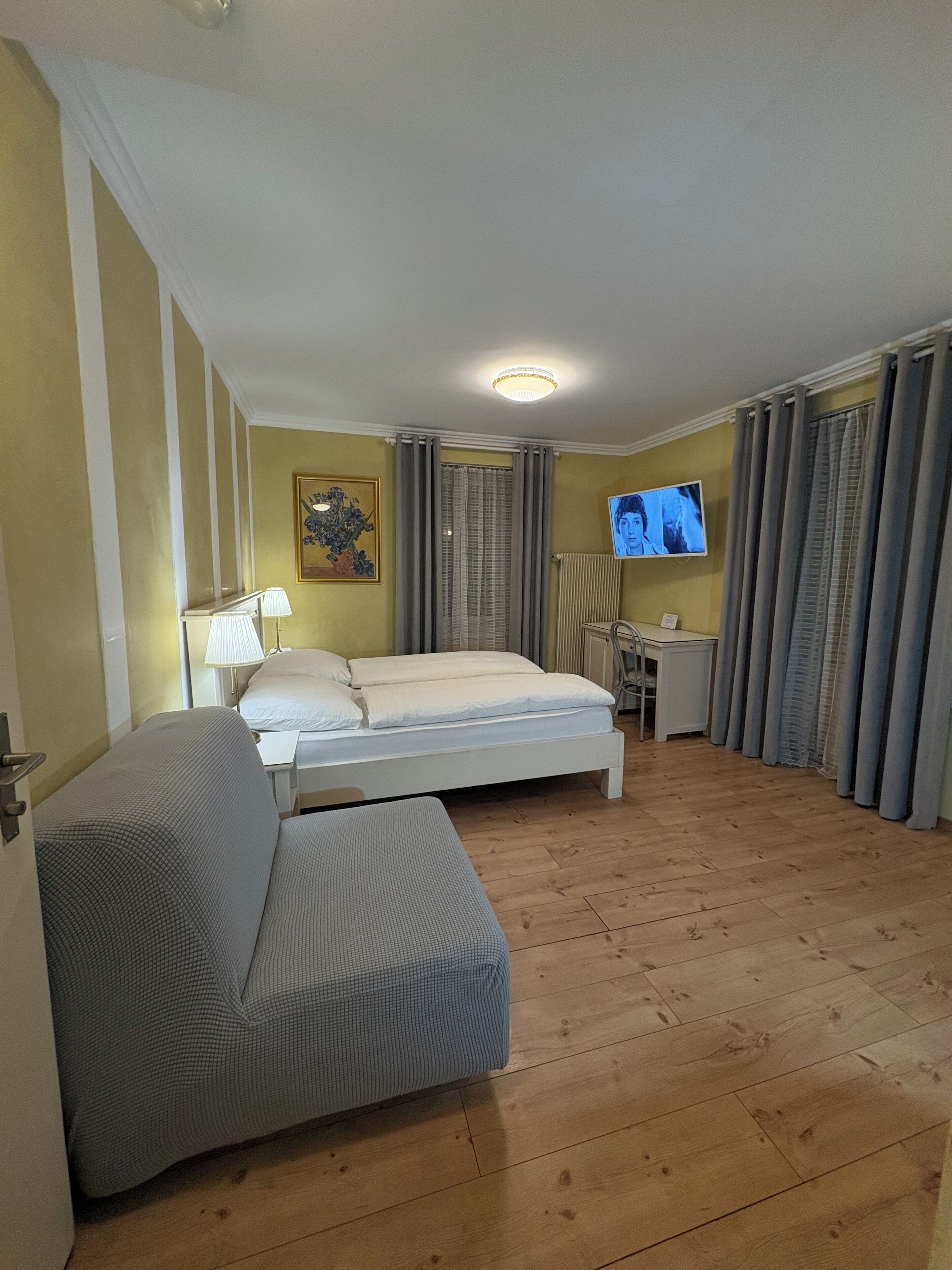 Quarto de hotel com cama, escrivaninha, cadeira, televisão e um quadro na parede.