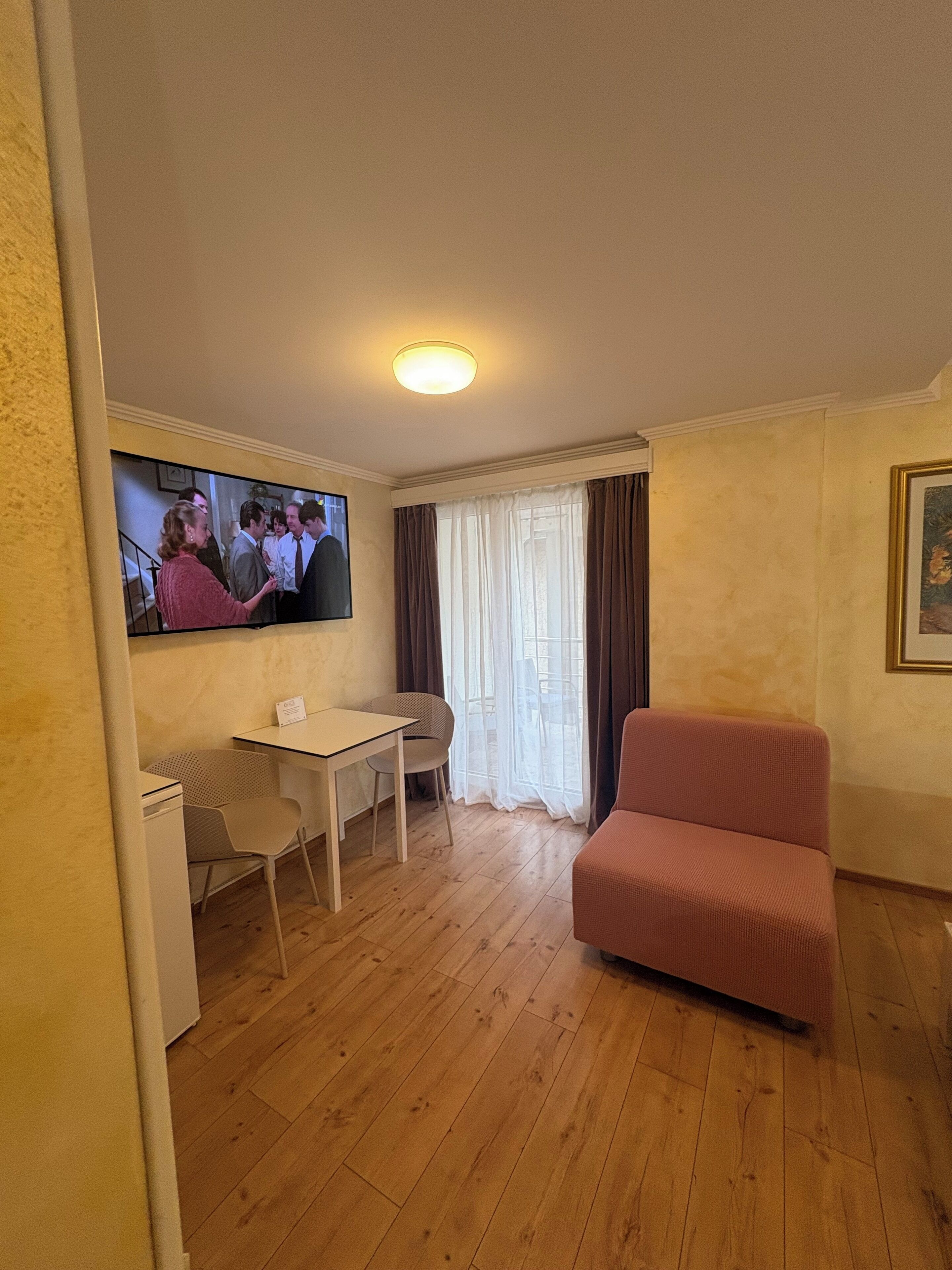 A living room with a television, a pink sofa, a dining table with chairs, and a window with curtains.