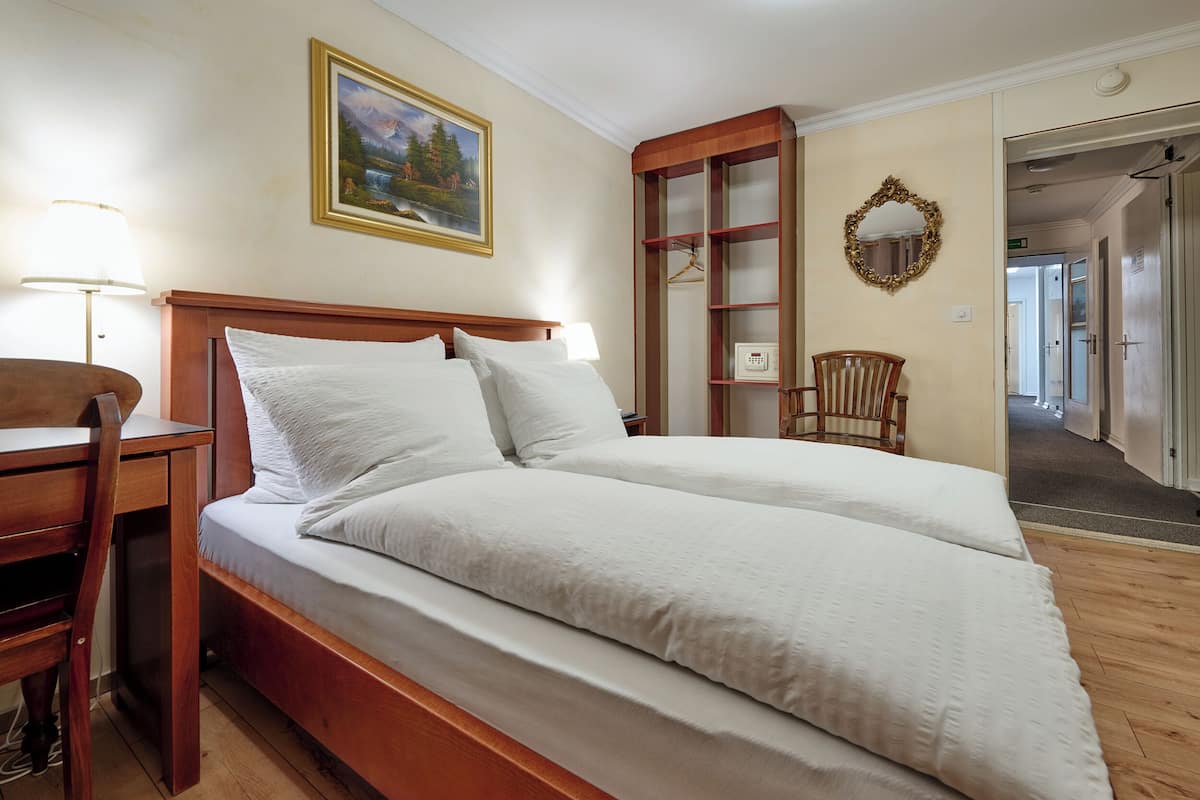 A double bed with white linens, a wooden desk with a lamp, a framed painting on the wall, a mirror, and a wooden chair.