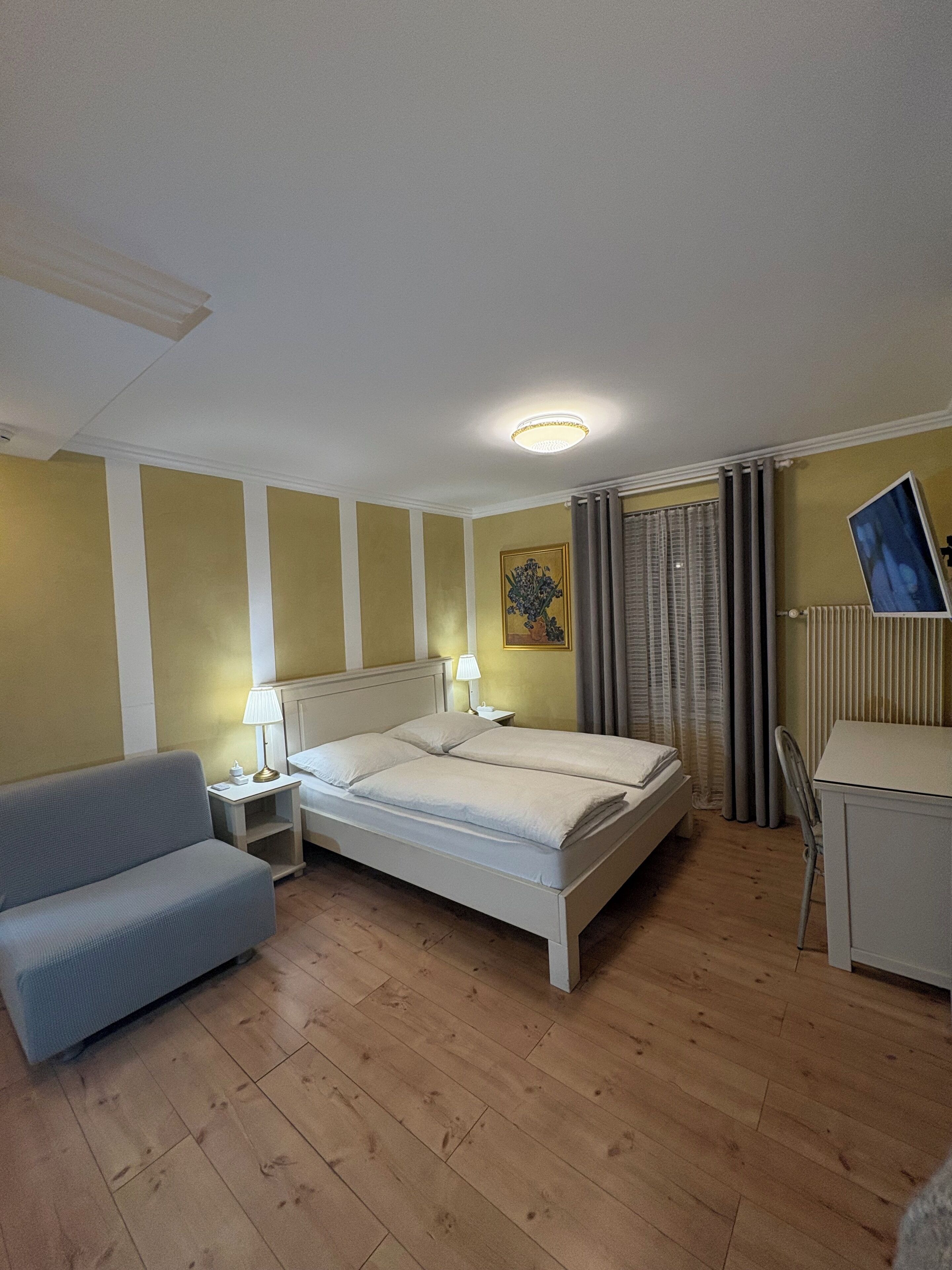 Habitación de hotel con cama, escritorio, silla, mesita de noche, lámpara y un cuadro en la pared.