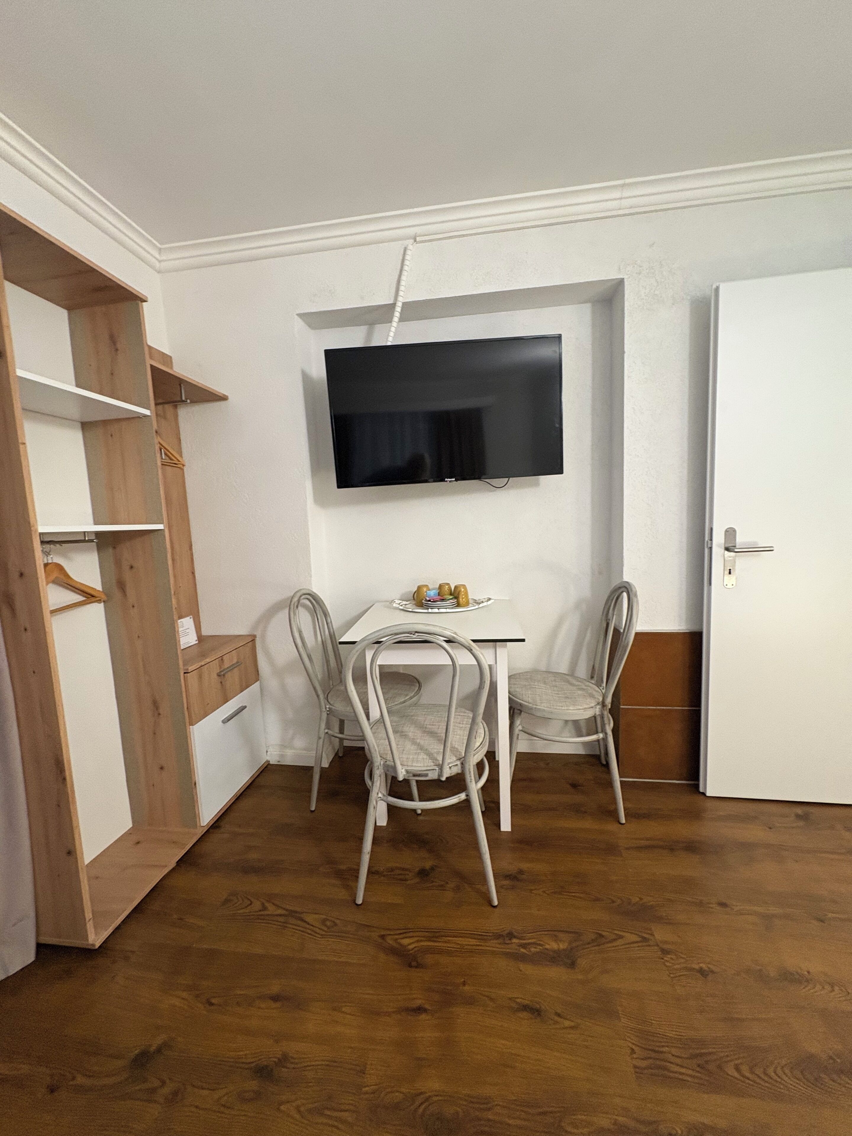 A small room with a white table, two chairs, a television mounted on the wall, and a wooden wardrobe.