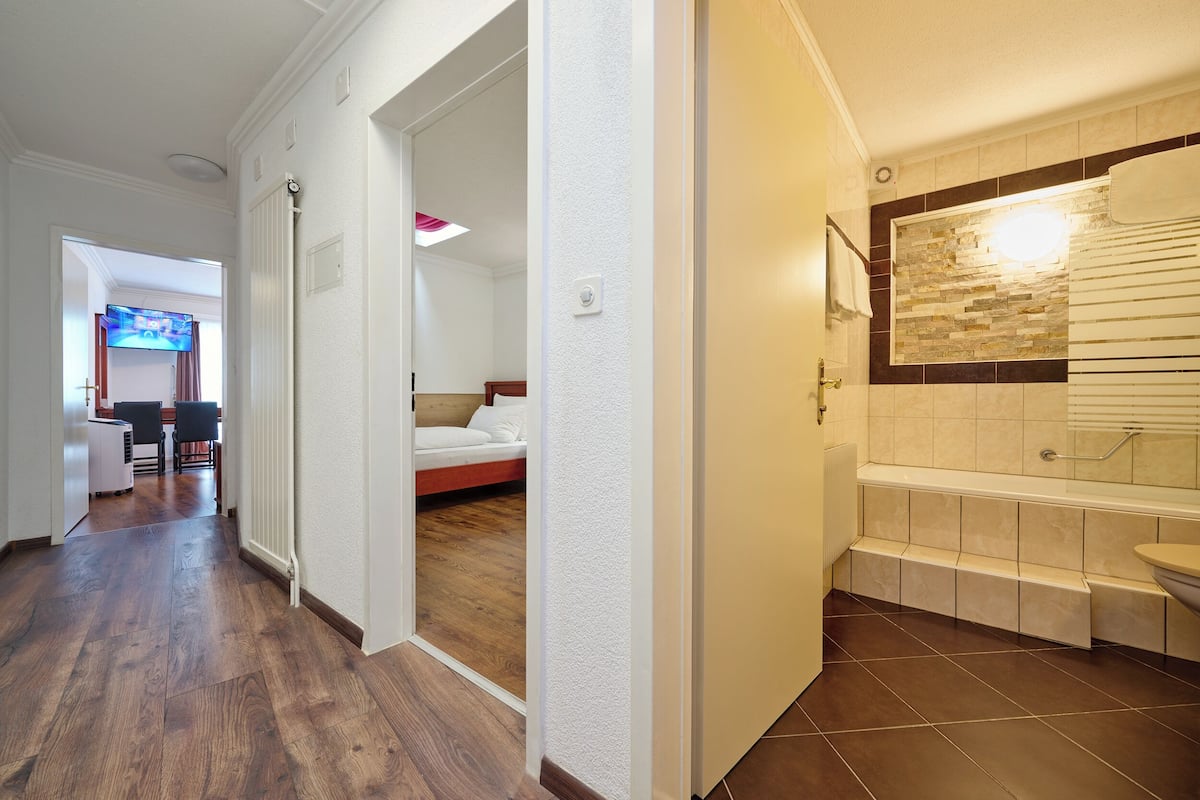 A hotel room with a wooden floor, a bed, a bathroom with a bathtub, and a TV in the living area.