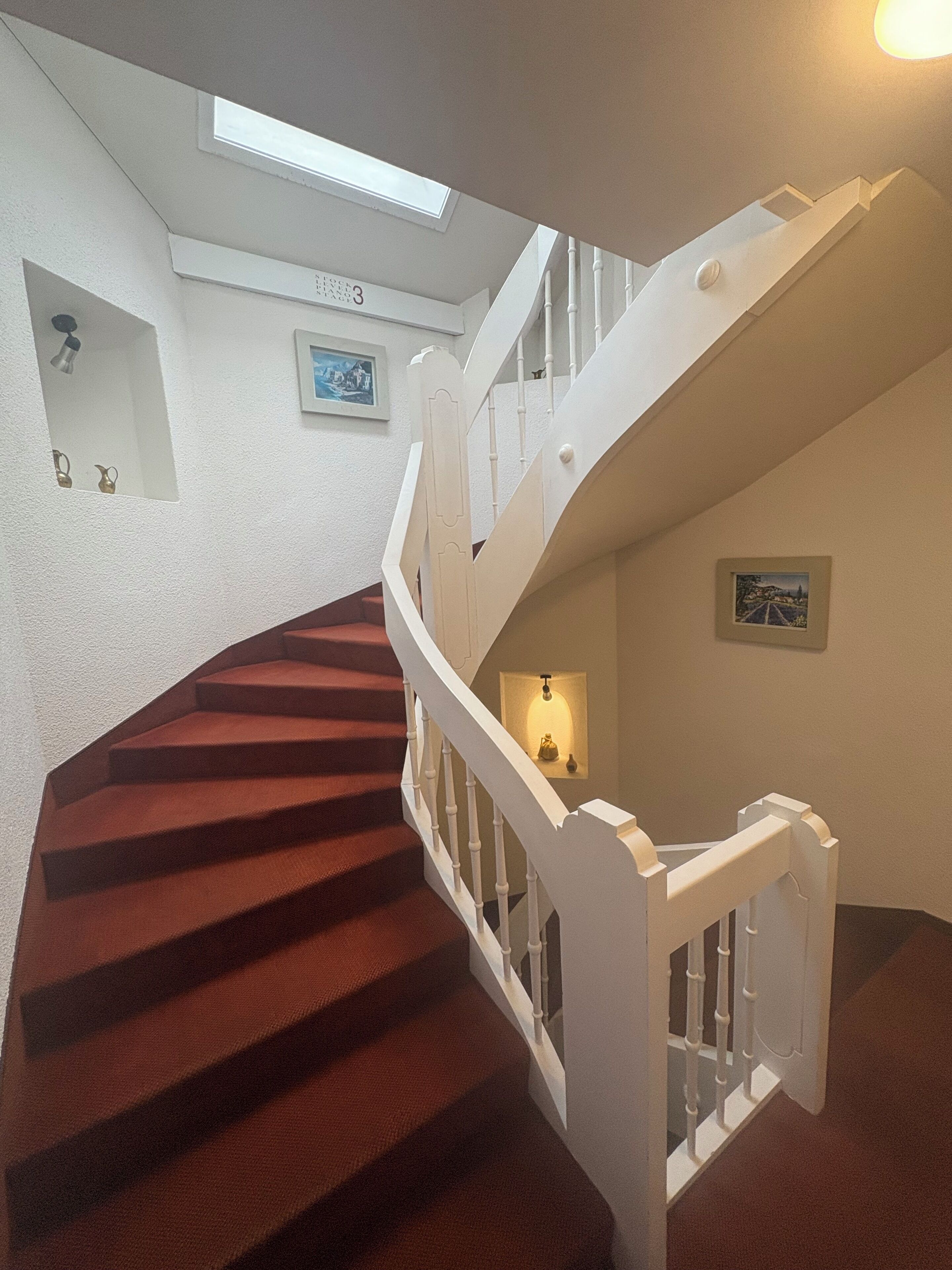 Un escalier avec une rampe blanche, une moquette rouge et un petit luminaire fixé au mur.
