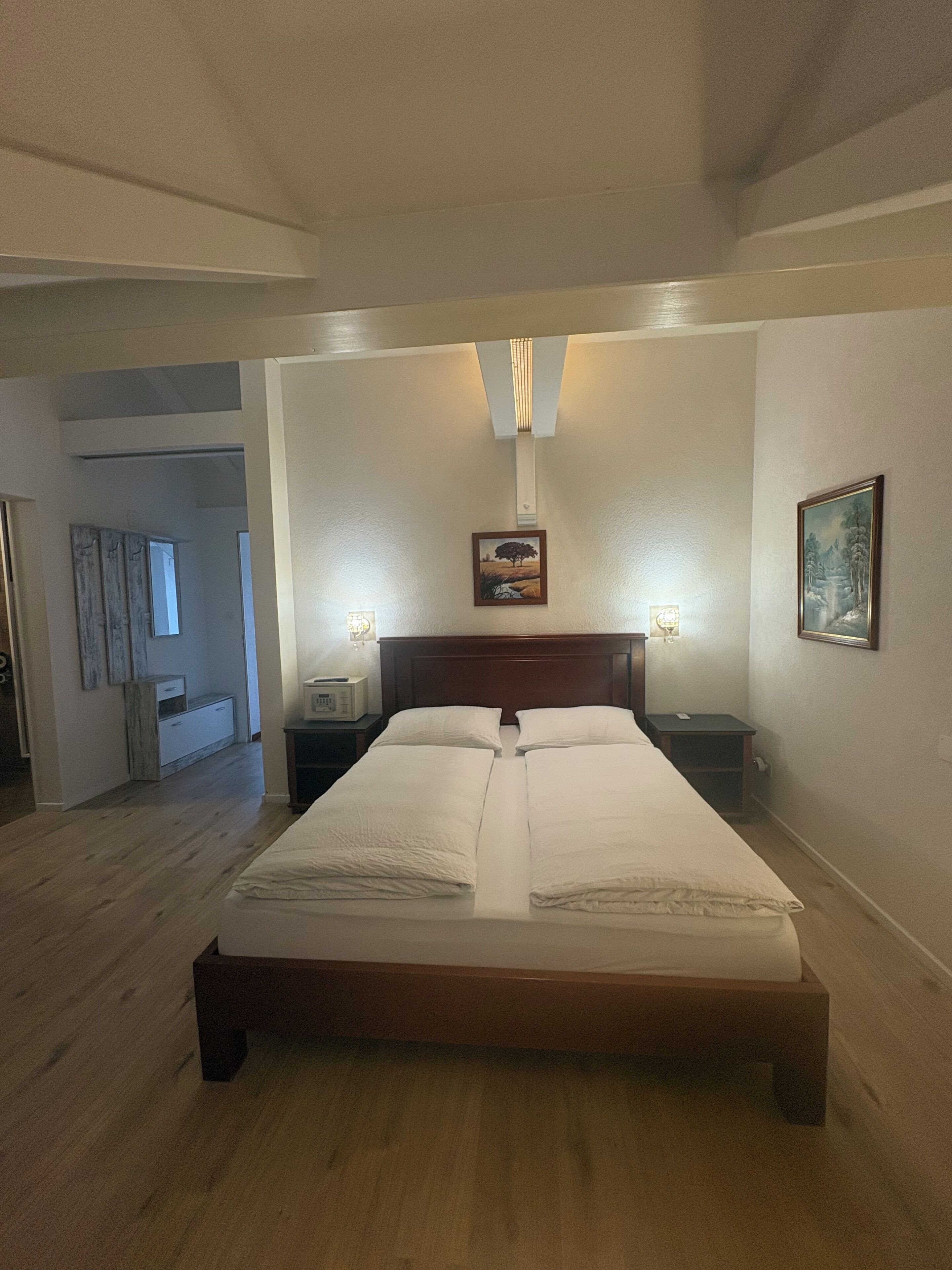 A bedroom with a large bed, two bedside tables, a nightstand, a wall-mounted lamp, and a painting.