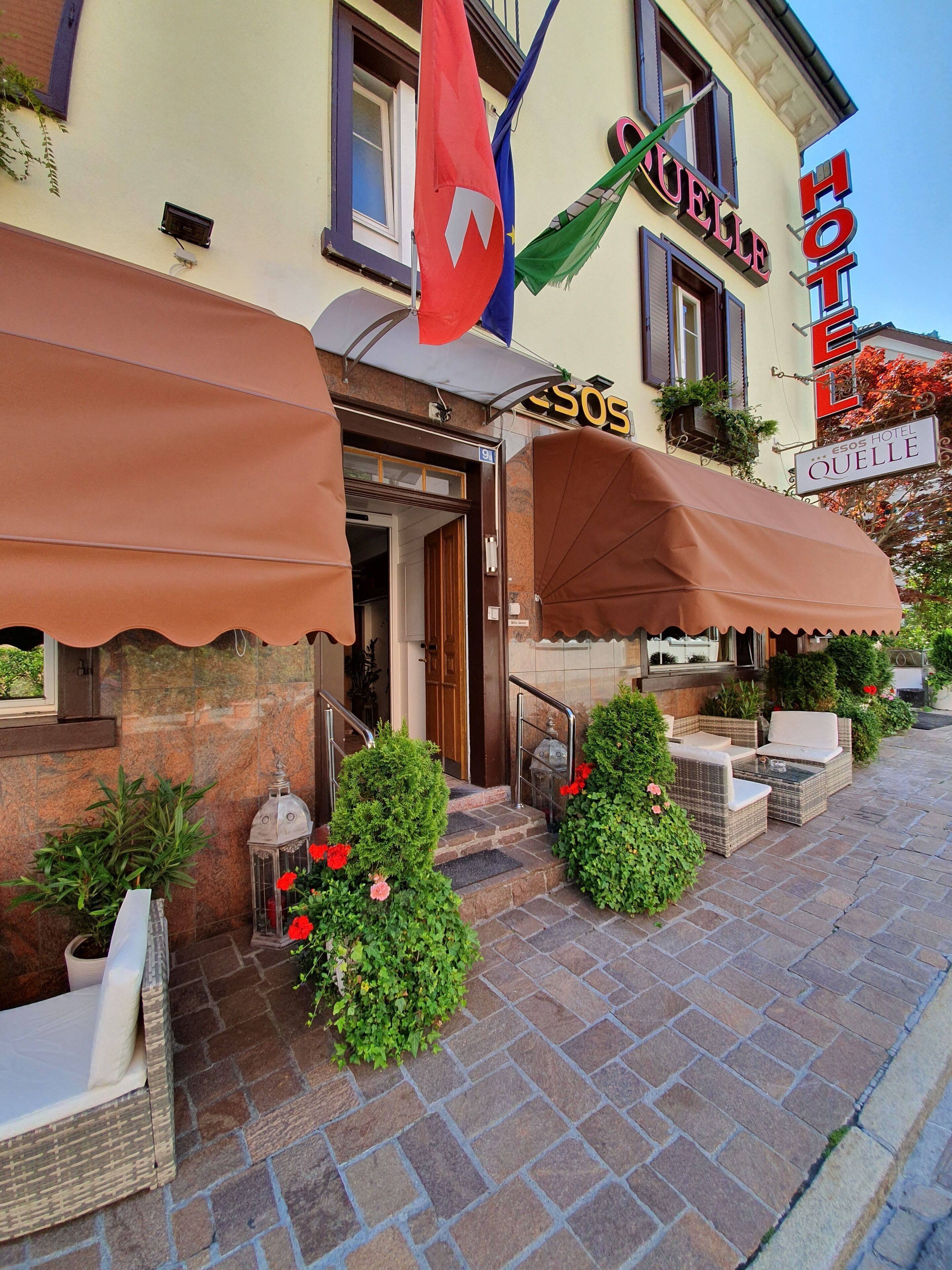 A hotel entrance with a covered outdoor seating area, a flag, and a sign that reads "Hotel Esos."