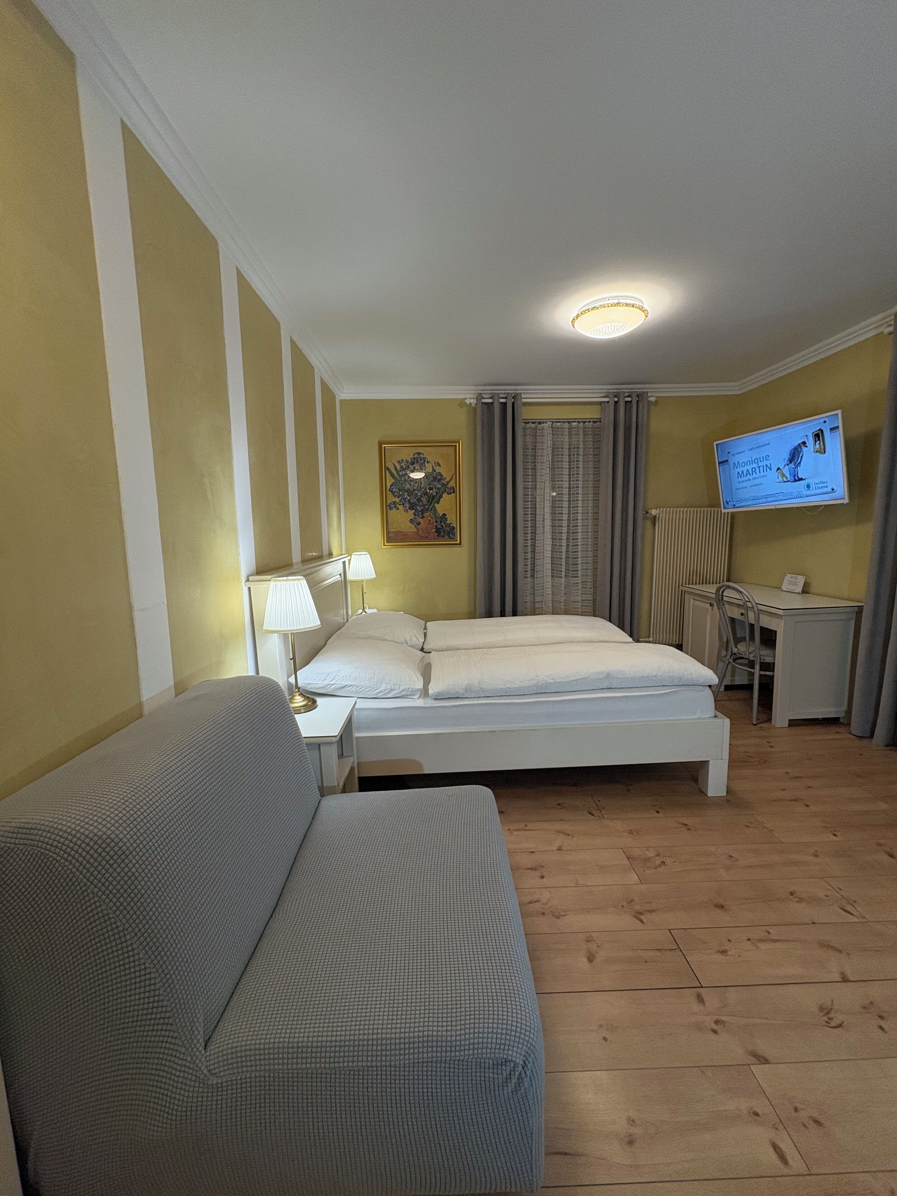 Una habitación de hotel con cama, sofá, escritorio, televisor y un cuadro en la pared.