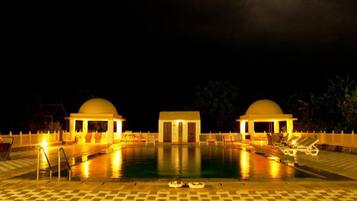 Un área de piscina nocturna con dos estructuras abovedadas, un edificio central y sillones alrededor de la piscina.