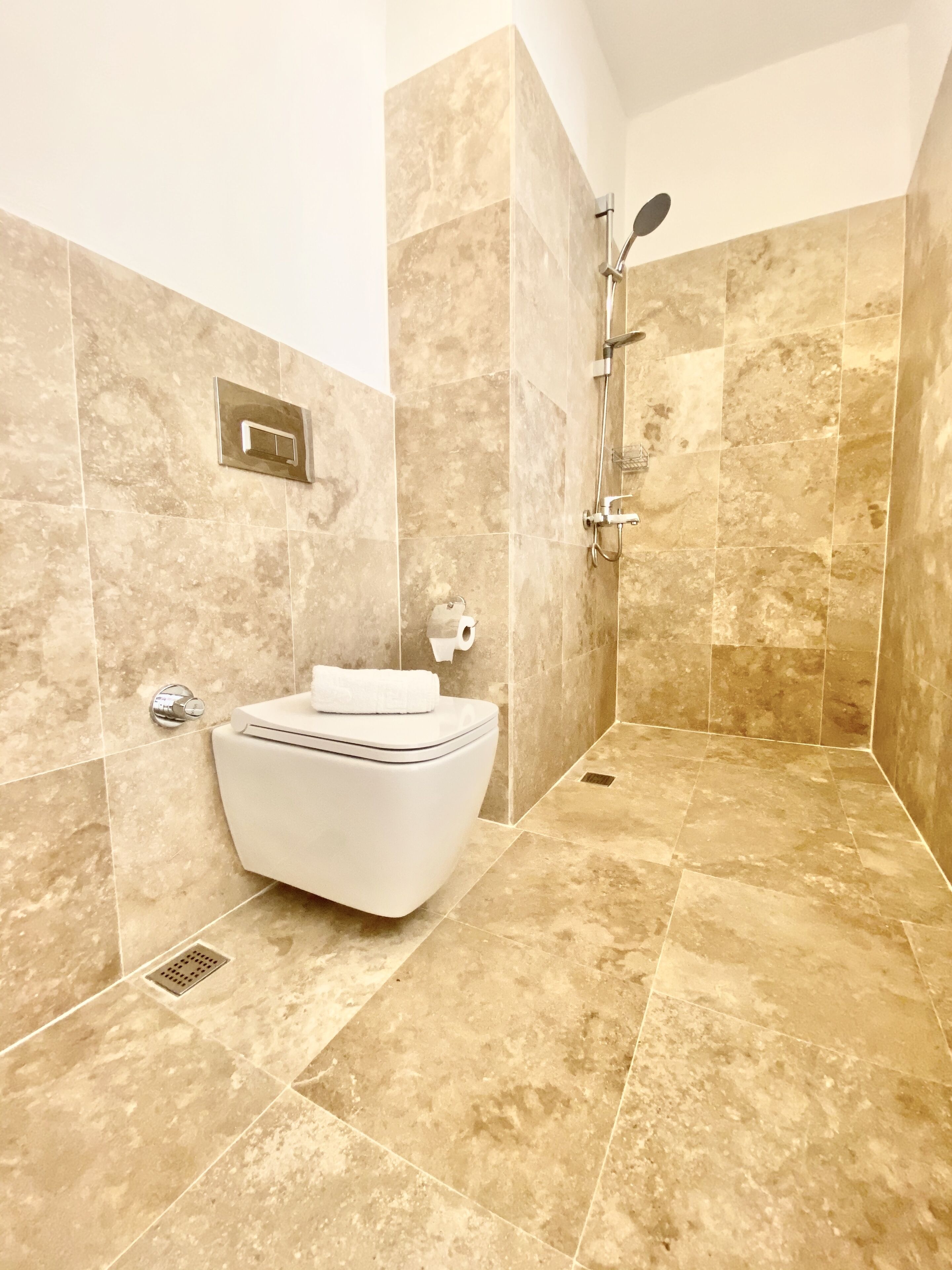 Et badeværelse med et hvidt toilet, en bruser med håndbruser og vægge beklædt med beige fliser.