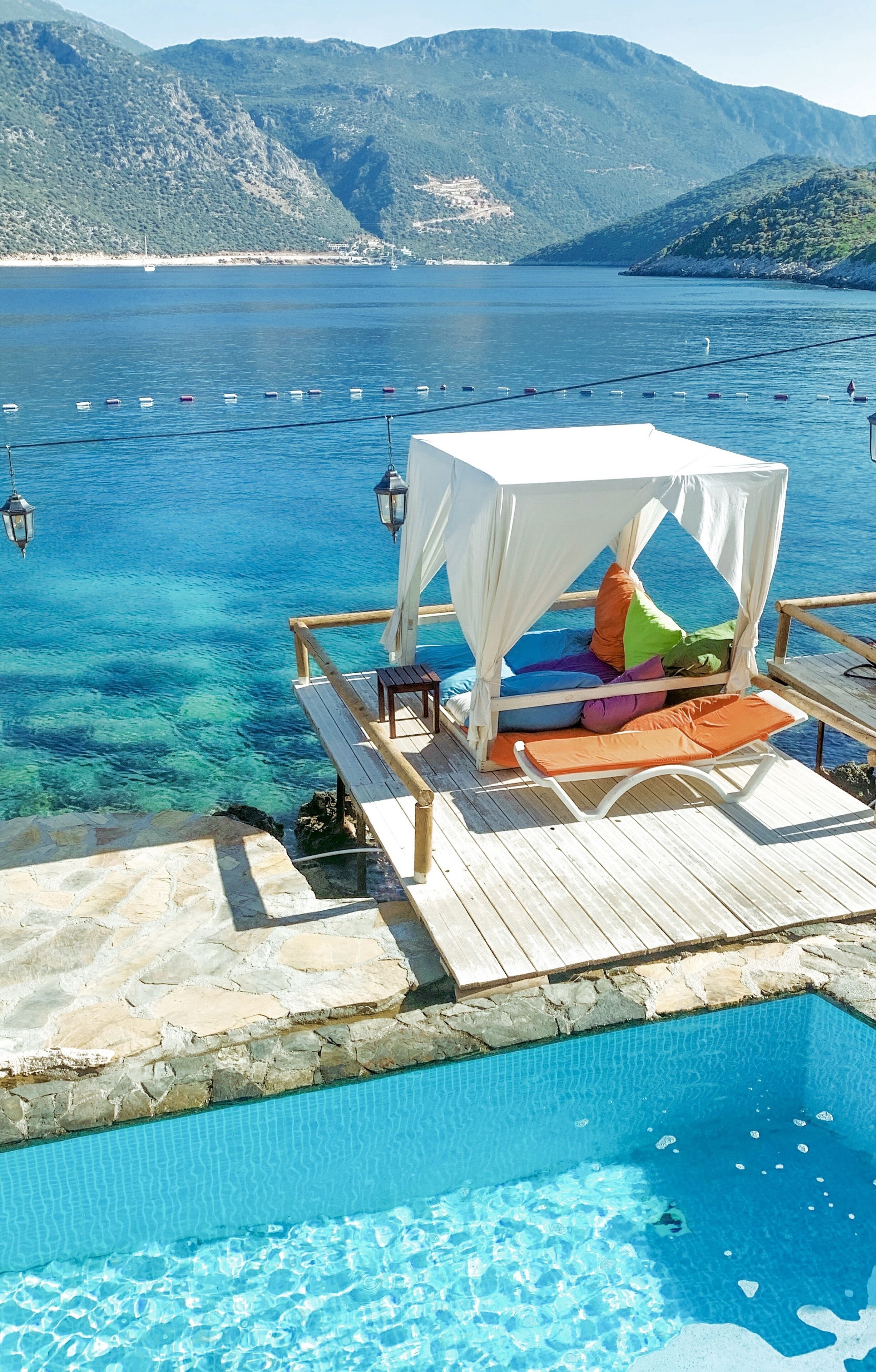 Zona junto a la piscina con una cama con dosel, agua azul cristalina y barcos al fondo.